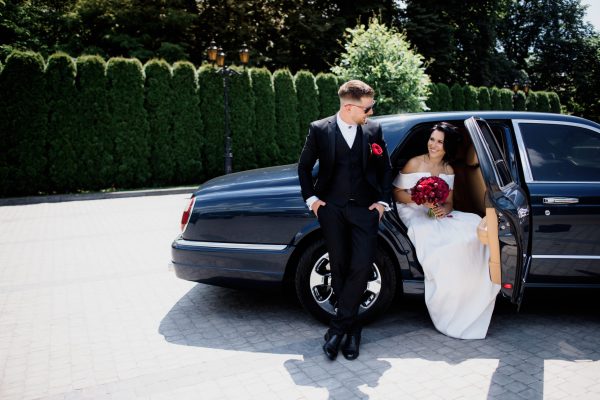 Beautiful wedding couple is smiling in the black car on the sunny day, dressed in elegant wedding outfits with red bouquet
